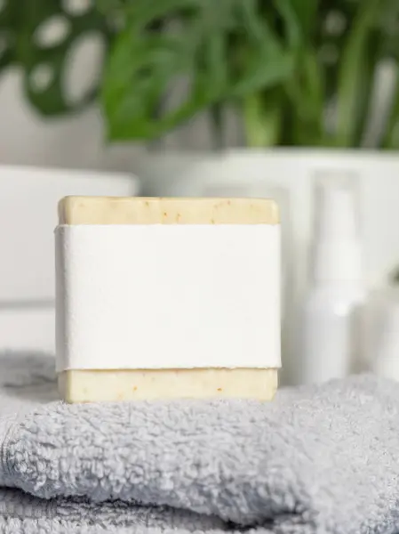 Beige handmade soap bar with blank label on light grey folded towel against basin and monstera plant close up, mockup.  Lifestile scene with hygiene and personal care products in bathroom
