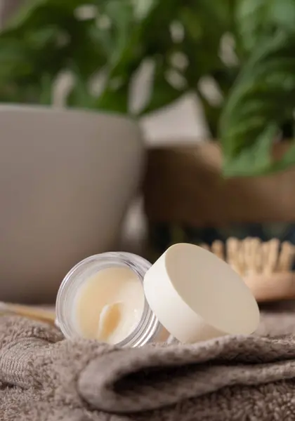 Opened lip balm jar with blank lid on beige folded towels near basin and green monstera, close up, cosmetic mockup.  Lifestile scene with skincare product in bathroom
