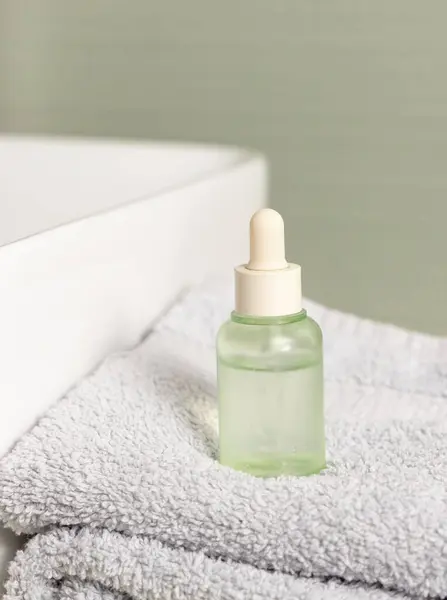 Green dropper bottle near basin on light grey towel against green wall in bath, close up. Natural personal care cosmetics, everydy female beauty routine