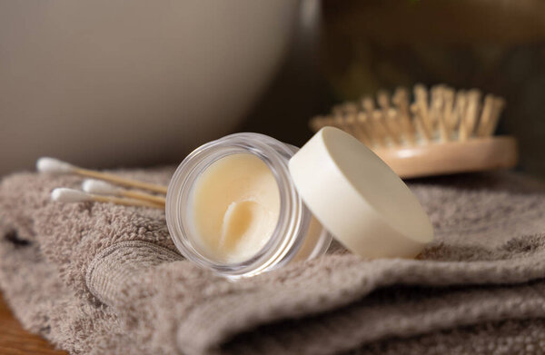 Открытая банка для бальзама для губ с пустой крышкой на бежевых сложенных полотенцах возле щетки и ватные тампоны против раковины, крупным планом, косметический макет. Lifelle сцена с продуктом Skincare в ванной