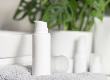 Beyaz olan, yeşil fabrikanın yanındaki gri katlanmış havluya kozmetik şişesi pompalıyor. Kapalı, kozmetik modelleme. Kadın kişisel bakım ürünleriyle modern yaşam sahnesi