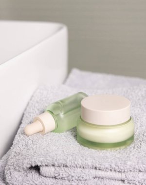Açık yeşil damlalık şişesi ve açık gri katlanmış banyo havlusuna kapalı krema kavanozu. Havuz yakın çekim, kozmetik model. Doğal organik ürünlerle güzellik ve cilt rutinleri