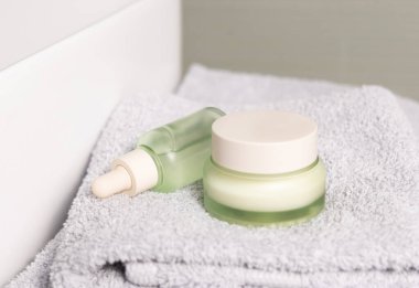 Açık yeşil damlalık şişesi ve açık gri katlanmış banyo havlusuna kapalı krema kavanozu. Havuz yakın çekim, kozmetik model. Doğal organik ürünlerle güzellik ve cilt rutinleri