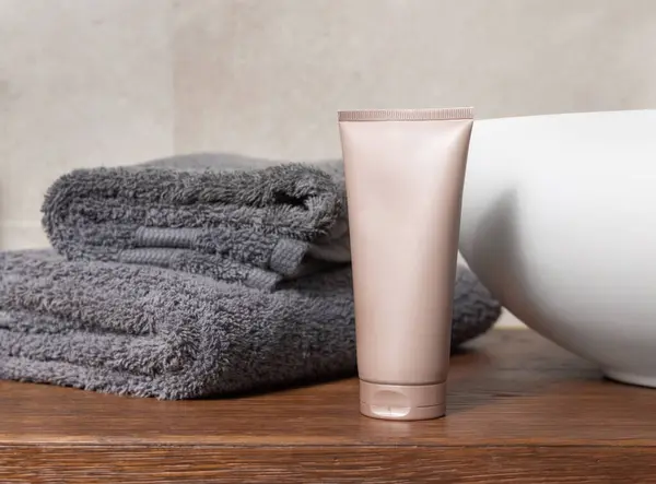 Beige cosmetic tube near grey folded towel on vessel basin on brown wooden countertop in beige bathroom, close up, cosmetic mockup for brand packaging
