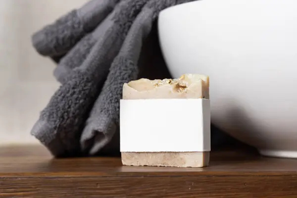 Soap bar with blank label near grey folded towel on vessel basin on brown wooden countertop in beige bathroom, close up, mockup for brand packaging. Natural handmade hygiene product