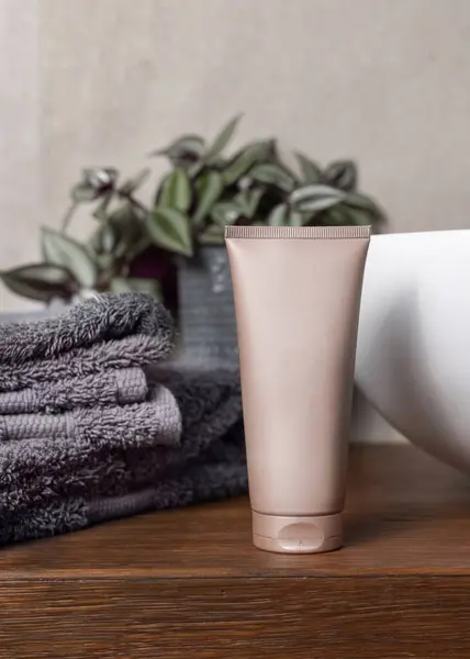 Beige cream squeeze tube near grey folded towels, green plant and vessel basin on brown wooden countertop in bathroom, close up, brand packaging mockup. Natural cosmetic product