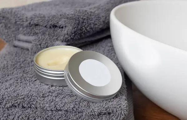 Stock image White opened body butter jar with label near dark grey folded towel and vessel basin on brown wooden countertop in bathroom, close up, cosmetic mockup for brand packaging