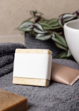 Koyu gri havluların üzerinde boş etiket olan sabun barı, banyoda yeşil bitki ve damar havzasının yanında, yakın plan, marka ambalajlama modeli. Erkekler için hijyen ürünü