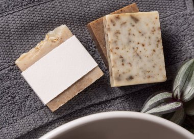 Gri katlanmış havlularda boş etiketli sabun barı, banyoda yeşil bitki ve damar havzasının yanında, yakın plan, marka ambalajlama modeli. Doğal el yapımı hijyen ürünü.