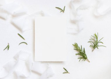 Beyaz ipek kurdelelerin yanındaki boş kart ve biberiye yaprakları üst manzara, düğün modeli. Dikey kartlı ve aromatik yapraklı romantik bir sahne. Sevgililer Günü, Bahar veya Anneler Günü konsepti