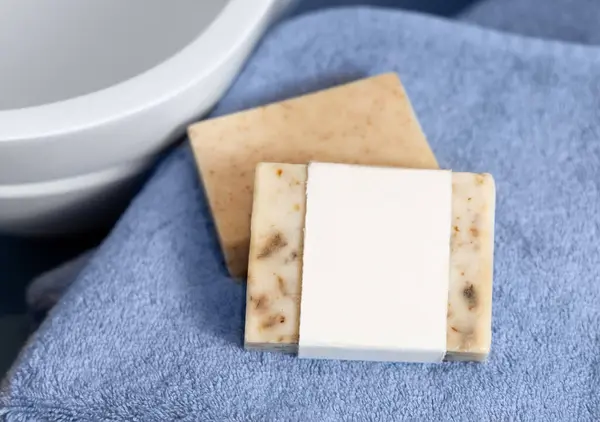 stock image Beige soap bar with blank label on blue towels near vessel sink in bathroom, close up, brand packaging mockup. Natural handmade hygiene product