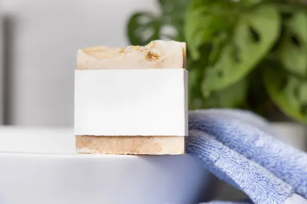 stock image Beige soap bar with blank label on vessel sink near blue towels and plant in bathroom, close up, brand packaging mockup. Natural handmade hygiene product