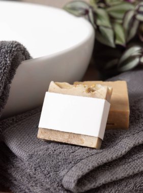 Koyu gri havluların üzerinde boş etiket olan sabun barı, banyoda yeşil bitki ve damar havzasının yanında, yakın plan, marka ambalajlama modeli. Erkekler için hijyen ürünü