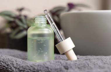 Pipette yeşil cam bir kozmetik şişeye kurulmuş. Koyu gri havluyu, leğene ve yeşil bitkiye sürmüş. Yakın plan. Erkekler cilt bakımı veya sağlık ürünleri, yağ veya seru