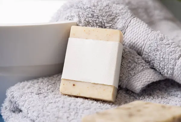 stock image Beige soap bar with horizontal blank label on light grey towels near vessel sink in bathroom, close up, brand packaging mockup. Natural handmade hygiene product