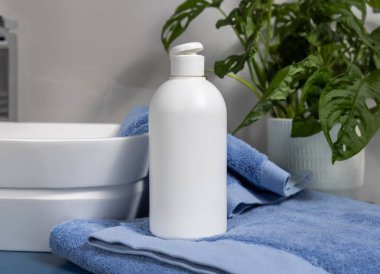 Banyodaki yeşil bitki ve damar havuzu yakınındaki mavi katlanmış havluların üzerinde beyaz, boş kozmetik kapak kapağı. Kapalı, marka ambalaj modeli. Doğal losyon, sabun ya da şampuan.