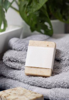 Damar lavabosunun yanında açık gri havlular üzerinde boş etiketli bej sabun barı ve banyoda yeşil bitki, yakın plan, marka ambalaj modeli. Doğal el yapımı hijyen ürünü.