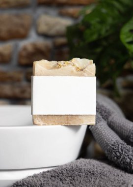 Taşıyıcı üzerinde boş etiketi olan bej sabun, taştan duvara karşı koyu gri havlular ve banyoda yeşil bitki, yakın plan, marka ambalaj modeli. Doğal el yapımı hijyen ürünü.