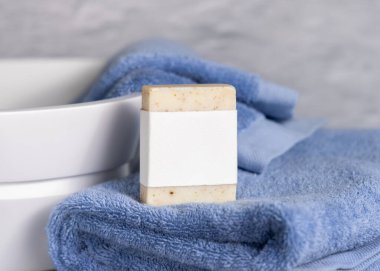 Mavi katlanmış havluların üzerinde boş etiketi olan bej sabun barı banyoda lavabonun yanında, yakın plan, marka ambalaj modeli. Kişisel bakım için doğal el yapımı hijyen ürünü.