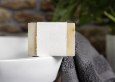 Taşıyıcı üzerinde boş etiketi olan bej sabun, taştan duvara karşı koyu gri havlular ve banyoda yeşil bitki, yakın plan, marka ambalaj modeli. Doğal el yapımı hijyen ürünü.