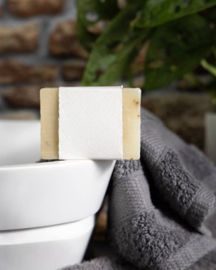 Taşıyıcı üzerinde boş etiketi olan bej sabun, taştan duvara karşı koyu gri havlular ve banyoda yeşil bitki, yakın plan, marka ambalaj modeli. Doğal el yapımı hijyen ürünü.