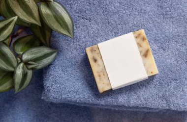 Yeşil fabrikanın yanındaki mavi katlanmış havluların üzerinde boş etiketli bej sabun barı, üst görünüm, marka ambalaj modeli. Doğal el yapımı hijyen ürünü.