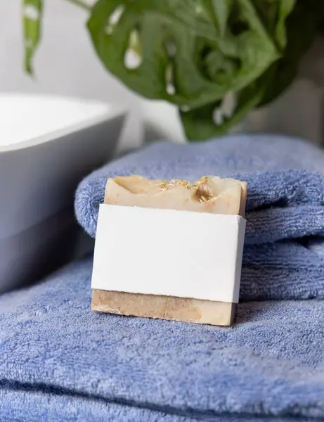 stock image Beige soap bar with blank label on blue folded towels near vessel sink and green plant in bathroom, close up, brand packaging mockup. Natural handmade hygiene product