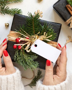 Yatay hediye etiketi, köknar ağacı dalları, çam kozalakları ve Noel dekoru olan elleri beyaz masanın üstünde. Boş etiketli kış kompozisyonu Mockup, boşluğu kopyala
