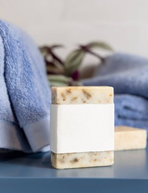 Mavi katlanmış havluların yanında boş etiketli bej sabun barı ve banyoda yeşil bitki, yakın plan, marka ambalaj modeli. Doğal el yapımı hijyen ürünü.