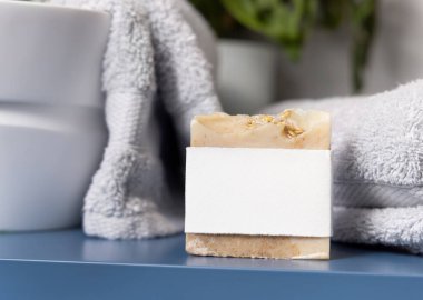 Açık gri havluların yanında boş etiketli bej sabun barı, mavi banyoda damar lavabosu ve yeşil bitki, yakın plan, marka ambalaj modeli. Doğal el yapımı hijyen ürünü.