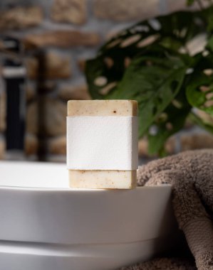 Taşıyıcı üzerinde boş etiketi olan bej sabun, taştan duvara karşı koyu gri havlular ve banyoda yeşil bitki, yakın plan, marka ambalaj modeli. Doğal hijyen ürünü.