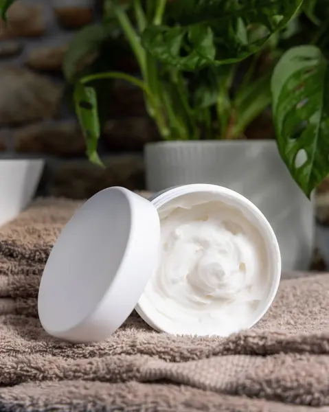 Stock image Opened cream jar with blank lid on brown folded towel near green plant in bathroom close up, skincare cosmetic packaging mockup.  Facial care for men