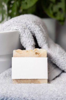 Damar lavabosunun yanında açık gri havlular üzerinde boş etiketli bej sabun barı ve banyoda yeşil bitki, yakın plan, marka ambalaj modeli. Doğal el yapımı hijyen ürünü.
