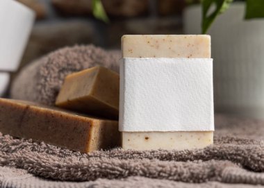 Açık kahverengi havluların üzerinde açık kahverengi etiketli bej sabun barı ve banyoda yeşil bitki, yakın plan, ambalaj modeli. Doğal el yapımı hijyen ürünü.