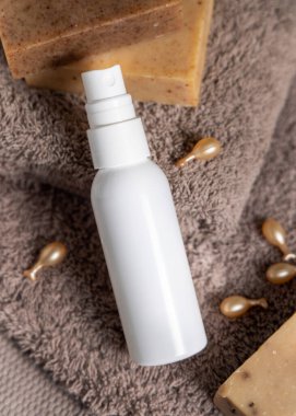 Beyaz sprey şişesi, sabun barları ve kozmetik kapsüllerin yanındaki kahverengi katlanmış havluların üzerinde. Üst görünüm, ambalaj modeli. Erkeklerin cilt bakımı için doğal kozmetik ürün veya deodorant