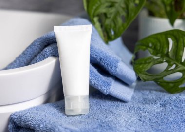 Küvetin yanında mavi banyo havlularının üzerinde beyaz krem sıkma tüpü ve banyoda yeşil bitki, yakın çekim, ambalaj modeli. Cilt bakımı için doğal kozmetik ürünü