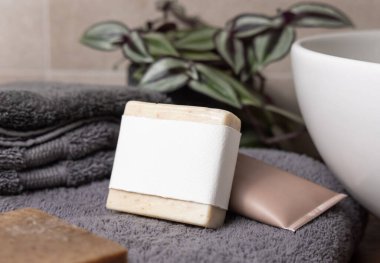 Gri katlanmış havluların yanında boş etiketli bej sabun barı, banyoda yeşil bitki ve damar havuzu, yakın plan, marka ambalajlama modeli. Doğal el yapımı hijyen ürünü.