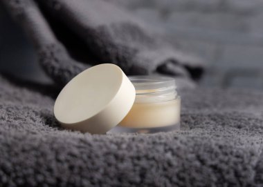 Banyoda katlanmış koyu gri havlunun üzerinde boş kapaklı dudak kremi kavanozu, yakın plan, ambalaj modeli. Erkeklerin kişisel bakımı için doğal el yapımı kozmetik ürünler