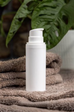 Banyoda yeşil fabrikanın yanındaki kahverengi havluların üzerinde beyaz krem pompa şişesi, yakın çekim, kozmetik ambalaj modeli. Kişisel bakım için doğal erkek yüz kremi