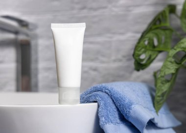 White cream squeeze tube on vessel sink near blue towels and green plant in bathroom, close up, packaging mockup. Natural cosmetic product for face or body care clipart