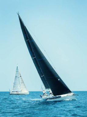 Mavi bulutsuz gökyüzünün altında yelkenli yarışı yapar. Mürettebat güvertede birlikte çalışıyor, diğer yelkenli arka planda görünüyor. Denizcilik rekabetinin ve takım çalışmasının özü