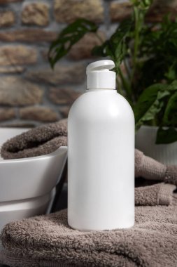 Endüstriyel banyodaki taş duvara karşı yeşil bitki ve kap kapaklarının yanındaki kahverengi havluların üzerinde beyaz boş kozmetik kapak şişesi, losyon, sabun ya da şampuan için yakın plan.