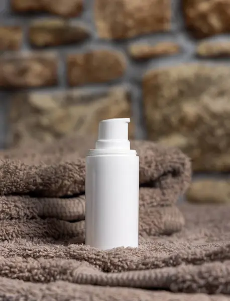 stock image White pump bottle on light brown folded towels against stone wall, close up, packaging mockup. Natural cosmetic product for face or body care cream or serum