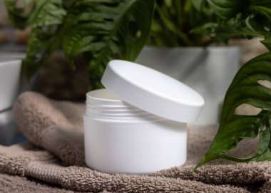 Banyoda yeşil bitkinin yanında kahverengi katlanmış havluya beyaz krema kavanozu aç, kapat. Yüz bakımı için konteynırda doğal kozmetik ürünler, rutin cilt bakımı.