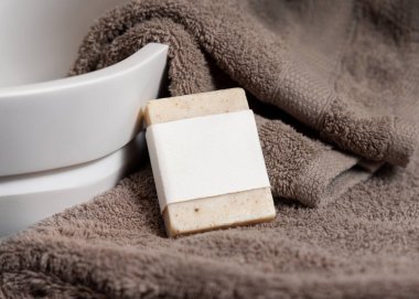 Damarın yanındaki kahverengi havluların üzerinde boş etiket olan bej sabun barı banyoda lavabo, kapanış, marka ambalaj modeli. Doğal el yapımı hijyen ürünü.