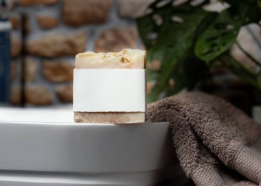 Damarında boş etiketi olan bej sabun çubuğu taştan duvara karşı kahverengi havluların yanında lavabo ve banyoda yeşil bitki, yakın plan, marka ambalaj modeli. Doğal hijyen ürünü.