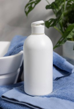 Banyodaki yeşil bitki ve damar havuzu yakınındaki mavi katlanmış havluların üzerinde beyaz, boş kozmetik kapak kapağı. Kapalı, marka ambalaj modeli. Doğal losyon, sabun ya da şampuan.