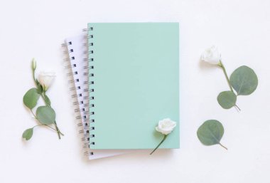 Yeşil okaliptüs yaprakları ve çiçeklerin üst görünüşü yakınındaki beyaz bir masanın üzerindeki açık yeşil kaplı spiral defter, boş ders kitabı modeli. Minimum okul yatağı. 