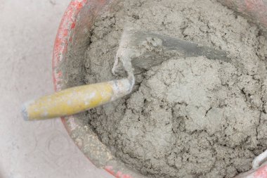 Kirli yüzeyde çimento ya da alçıyla kaplı bir kova. Etrafında çimento ve kum parçaları, içinde mala, çimento kalıntısı, üst manzara. İnşaat veya yenileme 