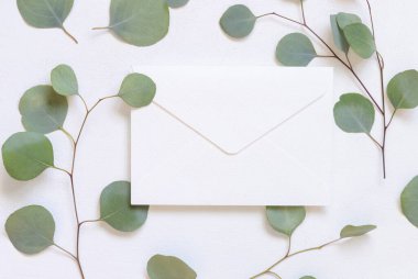 Yeşil okaliptüs dalları ve yaprakların yanında beyaz bir masanın üzerinde zarf, Mockup. Minimum romantik daire ve boş yatay kart. Düğün ve parti kırtasiyesi